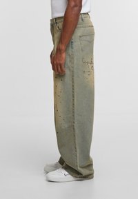 Muškarac nosi široke, izblijedjele zelene traperice s crnim mrljama od boje i bijele tenisice na jednostavnoj svijetloj pozadini.