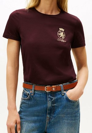 Mujer que lleva una camiseta de color púrpura oscuro con un emblema dorado y texto, combinada con vaqueros azules y un cinturón de cuero marrón con hebilla dorada.