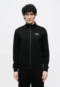 Mężczyzna o kręconych, ciemnych włosach ubrany w czarną kurtkę na zamek z logo EA7 Emporio Armani na piersi, stojący na jednolitym białym tle.