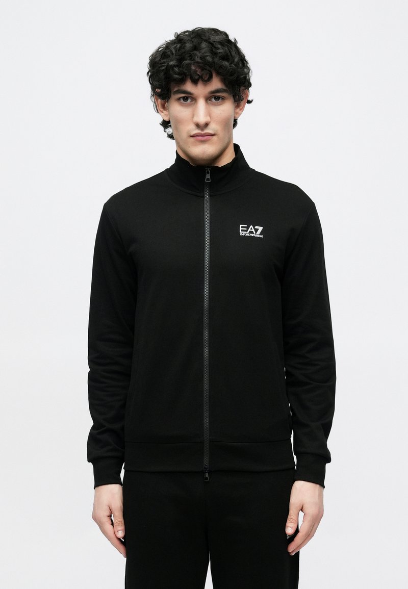 Mężczyzna o kręconych, ciemnych włosach ubrany w czarną kurtkę na zamek z logo EA7 Emporio Armani na piersi, stojący na jednolitym białym tle.