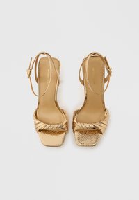 Χρυσά ψηλοτάκουνα σανδάλια με λουριά, με σχεδιασμό στριφογυρισμένου λουριού, τετράγωνη μύτη και υφή φινιρίσματος. Περιλαμβάνεται ρυθμιζόμενο κούμπωμα στον αστράγαλο.