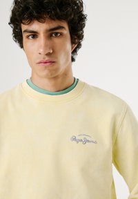 Geel katoenen sweatshirt met een ronde hals, voorzien van een blauw geborduurd logo. Gelaagd over een groene shirt met een textuur.