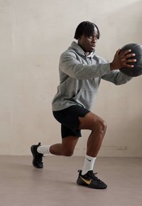 En atletisk mand i en grå hættetrøje og sorte shorts udfører et udfald, mens han holder en sort basketball. Han har sorte og gyldne sneakers og hvide sokker.