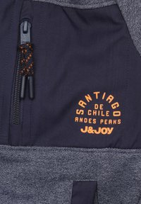 Marineblauwe en grijze jas van een gestructureerde stof. Bevat een oranje logo "Santiago de Chile Andes Peaks" en een ritszak met een zwarte trekband.