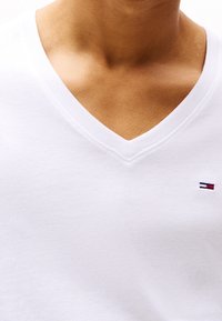 Primer plano del cuello y parte superior del pecho de una persona que lleva una camiseta blanca con cuello en V y un pequeño logo rojo, blanco y azul en el lado derecho.