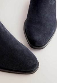 Stivaletti alla caviglia in suede blu navy con punta affilata, caratterizzati da una texture liscia e da una sottile suola nera, che evidenziano linee pulite e un design elegante.