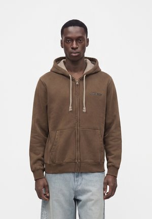 Ung mand iført en brun hættetrøje med lynlås og snore samt lyseblå jeans, stående mod en ensfarvet hvid baggrund.