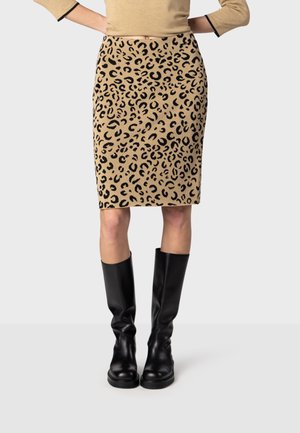 Femme portant une jupe beige et noire à motif léopard, un top beige et des bottes noires montantes, debout devant un fond clair uni.