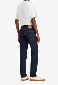 Mörkblå denimjeans med en texturerad yta, knappstängning, bakfickor och en ljusbrun läderlapp på midjan. Kombination med en vit T-shirt.