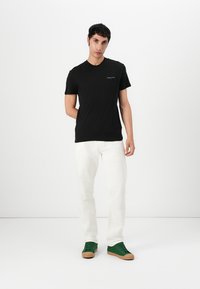 Fekete, rövid ujjú póló, amelyen egy kis logó található a mellrészen, fehér, slim-fit farmernadrággal és zöld, gumi talpú tornacipővel párosítva.