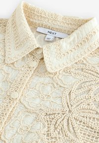 Blouse en dentelle crème avec broderie florale, boutons crème et col pointu, étiquetée avec les marques "NEXT" et "Made in China."