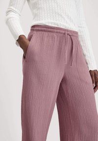 Nahaufnahme einer Person, die strukturierte mauvefarbene Jogginghosen und ein weißes Langarmoberteil trägt, mit Händen in den Taschen vor einem schlichten Hintergrund.