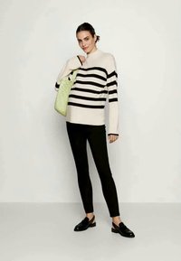 Sweter w paski w kolorze kremowym i czarnym, zestawiony z czarnymi legginsami i czarnymi mokasynami. Modelka trzyma torebkę o szorstkiej fakturze w zielonym kolorze.