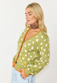 Ξανθιά γυναίκα φορώντας ελαφρύ πράσινο πουκαμίσα με πουά και πορτοκαλί περίγραμμα, καφέ κασκόλ με σχέδια στο λαιμό, χρυσούς κρίκους σκουλαρίκια και ανοιχτό μπλε τζιν.