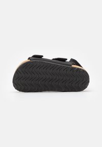 Svart sandal med en teksturert gummisåle, justerbare borrelåsremmer og en korkfotseng. Har et bølgete mønster på sålen for bedre grep.
