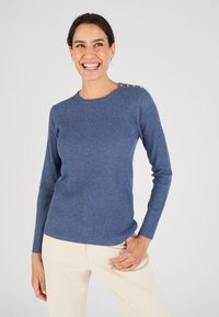 Pull à manches longues bleu côtelé avec un col rond et des accents de boutons décoratifs sur l'épaule, associé à un pantalon de couleur claire.