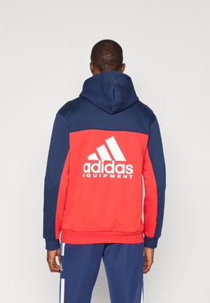 Man draagt een marineblauwe en rode Adidas hoodie met capuchon omhoog, van achteren, met het witte Adidas Equipment-logo op de rug.
