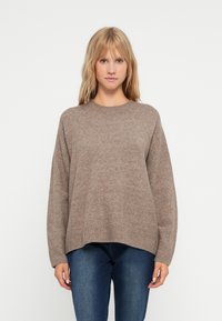Beige oversized genser med rund hals, ribbestrikket kant og lange ermer, laget av myk teksturert stoff. Sammen med blå jeans.
