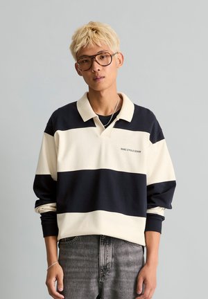Jeune personne aux cheveux courts décolorés porte de grandes lunettes, un t-shirt rayé noir et blanc à manches longues et un jean gris, debout devant un fond uni.