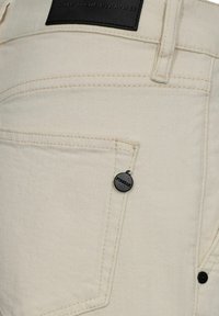 I pantaloni in denim beige presentano una texture sottile, cuciture a contrasto e una piccola etichetta nera con il marchio sulla tasca anteriore.