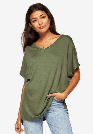 Oliwkowy, oversize T-shirt w serek z miękkiego materiału, charakteryzujący się luźnym krojem z bocznymi rozcięciami, zestawiony z jasnoniebieskimi dżinsami.