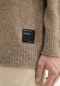 Beige gebreide trui met geribbelde textuur, voorzien van een zwart merklabel aan de zijkant. G toont over crèmekleurige broek.