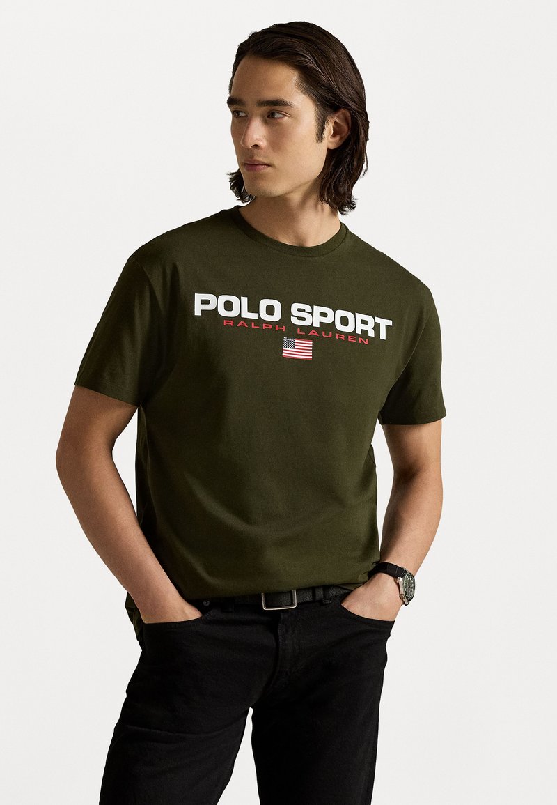 T-shirt en coton vert olive avec "POLO SPORT" et "RALPH LAUREN" imprimés en blanc et rouge. Présente un graphique de drapeau américain. Jean noir et montre.