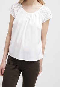 Haut blanc avec manches courtes en dentelle, encolure ronde et tissu froncé au niveau de la poitrine. Texture lisse avec ourlet incurvé. Associé à un pantalon foncé.