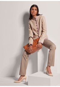 Beige strukturierte Jacke mit hohem Kragen, buntem Besatz, dunklen Knöpfen, kombiniert mit beigefarbenen Hosen und nudefarbenen High Heels; trägt eine braune Tasche.