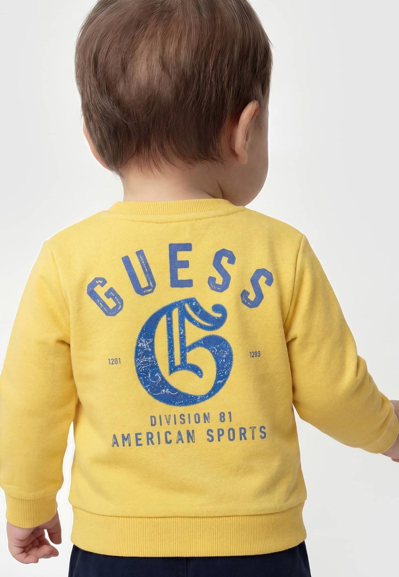 Enfant aux cheveux courts et bruns portant un sweat jaune avec un texte bleu "GUESS American Sports Division 81" dans le dos.