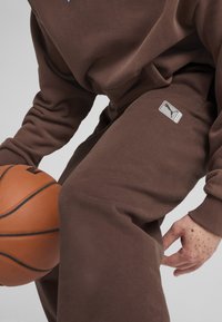 Hnedé tepláky z mäkkého materiálu s voľným strihom. Obsahujú malú nášivku loga Puma na boku a v ruke držiaceho basketbal.