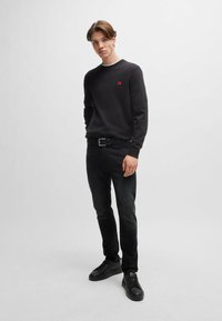Black crewneck sweatshirt with a small red logo, paired with dark jeans and black sneakers, featuring a smooth texture and fitted design.