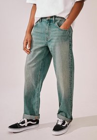 Jean en denim bleu clair usé avec une coupe ample, des ourlets roulés et des poches latérales, associés à des baskets noires et blanches.