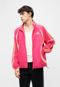 Młody mężczyzna w różowej sportowej kurtce Adidas z pomarańczowymi paskami, białym tank topem i czarnych spodniach, pozujący z jedną ręką w kieszeni.