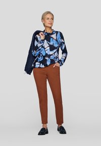 Langärmliges Oberteil mit floralem Muster in Blau und Weiß, kombiniert mit braunen, taillierten Hosen und schwarzen Slippern. Das Modell hält eine marineblaue Jacke.
