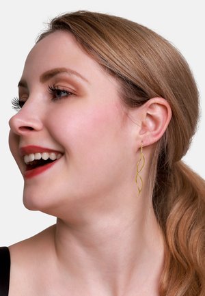 Orecchini spiralati color oro indossati da una donna. Il design presenta una forma slanciata e attorcigliata con una superficie liscia e lucida.