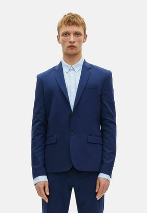 Blaues Sakko mit zwei vorderen Taschen, Ein-Knopf-Verschluss und Reverskragen. Darunter ist ein hellblaues Hemd mit Kentkragen.