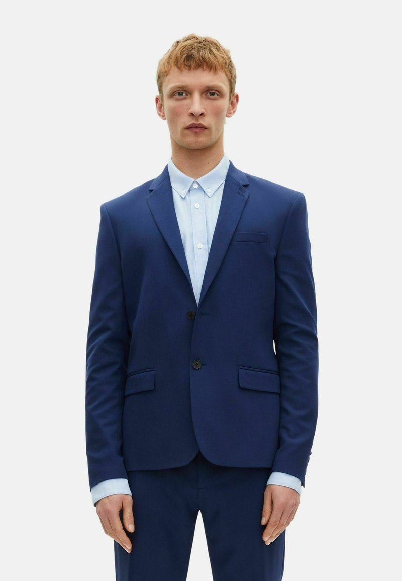 Blaues Sakko mit zwei vorderen Taschen, Ein-Knopf-Verschluss und Reverskragen. Darunter ist ein hellblaues Hemd mit Kentkragen.
