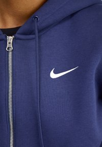 Laivastonsininen vetoketjuhuppari, joka on valmistettu pehmeästä kankaasta. Hupparissa on valkoinen Nike-logo rinnassa ja hopeiset vetoketjun osat.