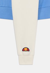 Manica di felpa bianca con polsino a coste blu. Presenta un logo ricamato rotondo con accenti rossi, gialli e blu navy vicino al polso.