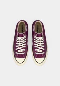 Violet aukštakulniai Converse sportbačiai su baltais raišteliais ir guminiais pirštų dangteliais, matomi iš viršaus ant balto fono.