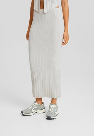 Kvinde iført en hvid ribstrikket midinederdel kombineret med lysegrå og hvide sneakers, stående på en ensartet lys baggrund.