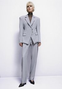 Άτομο με κοντά ξανθά μαλλιά που φοράει μια κομψή ανοιχτό γκρι παντελόνα, μαύρη διαφανή μπλούζα με υψηλό λαιμό και μαύρες ψηλοτάκουνες γόβες, στέκεται μπροστά σε λευκό φόντο.