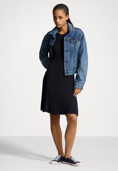 Femeie îmbrăcată într-o rochie neagră cu tricotaj în relief, o jachetă albastră din denim și adidași bleumarin, stând pe un fundal alb simplu.