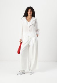 Blusa bianca con collo arricciato, maniche lunghe e bottoni frontali abbinata a pantaloni bianchi a gamba larga e borsa rossa. Sneakers in tela completano il look.