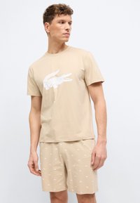 Άνδρας που φοράει μπεζ μπλουζάκι και ταιριαστά σορτς με λευκά λογότυπα κροκοδείλου, στέκεται και κοιτάζει πλάγια σε απλό φόντο.