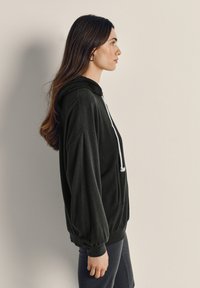 Žena s dlhými hnedými vlasmi, oblečená v čiernej mikine s bielymi šnúrkami a tmavými nohavicami, stojaca bokom na svetlom neutrálnom pozadí.