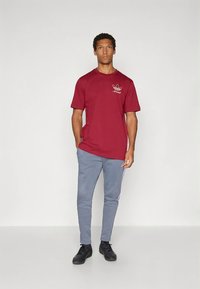 Raudonas medvilninis marškinėlis su apvalia iškirpte ir trumpomis rankovėmis, ant krūtinės esantis baltas Adidas logotipas. Derinamas su pilkomis siaurėjančiomis kelnėmis.