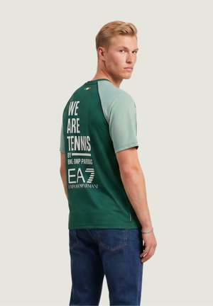 Młody mężczyzna z krótkimi blond włosami, ubrany w zielono-szarą koszulkę tenisową z napisem "WE ARE TENNIS" oraz niebieskie dżinsy, patrzący przez ramię.