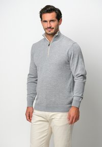 Grauer Strickpullover mit hohem Kragen und Reißverschluss, ausgestattet mit gerippten Bündchen und Saum. Getragen mit hellen Hosen für einen lässigen Look.